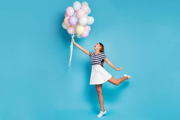 Full size profile photo of beautiful funky lady hold many colorful balloons fly up wind blowing wear striped t-shirt white short skirt footwear isolated blue color background - Фото, зображення