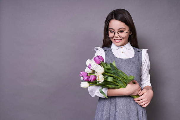 Young cute emotional child in glasses, holding multi-colored tulips in her hands and looking at them, happy with a gift to the left of the copy space, very joyful and surprised, on a gray background. - Fotoğraf, Görsel