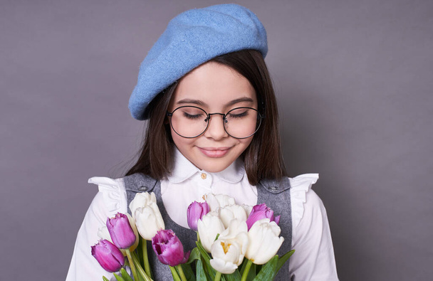 Cute young lady schoolgirl is a beautiful emotional long-haired brunette, in a stylish blue beret hat, in glasses, gently looks at flowers tulips in her hands, a French girl, on a gray background. - Фото, зображення