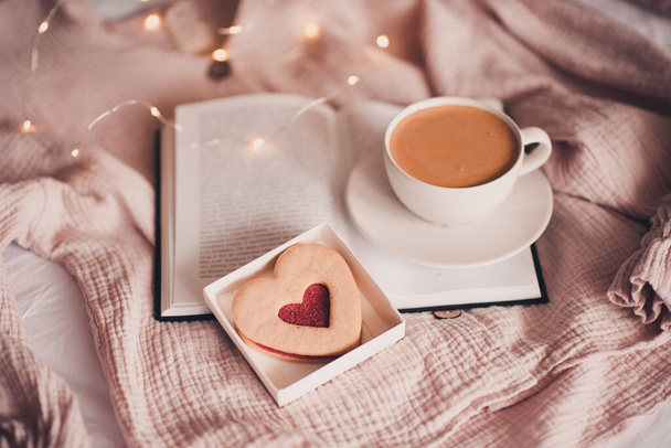 Frische Tasse Kaffee mit herzförmigem Kuchen auf aufgeschlagenem Buch im Bett über Lichtern Nahaufnahme. Guten Morgen. Frühstück. Valentinstag.  - Foto, Bild