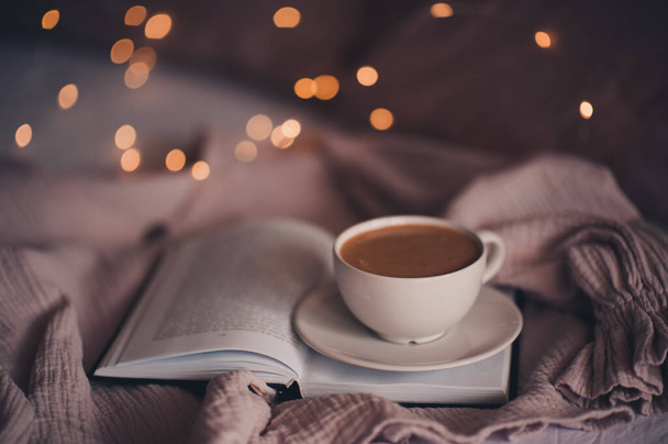 Cup of tasty coffee staying on paper book in bed over glowing lights closeup. Good morning. Breakfast. Winter season.  - Photo, Image