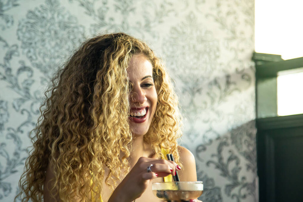 Woman with curly hair, a yellow dress and red painted lips laughing with a cocktail glass in her hand in front of a wall with vintage decoration - Photo, Image