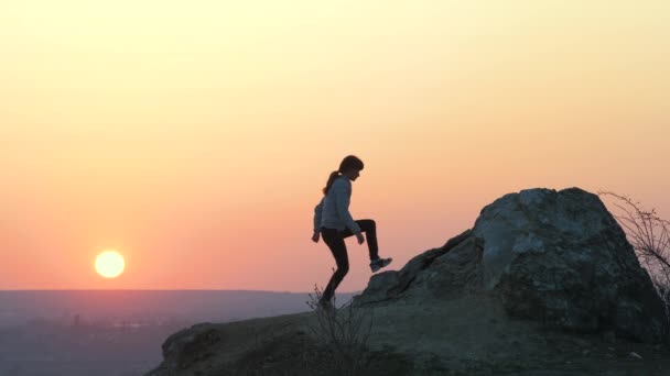 Gün batımında dağlarda tek başına büyük taşlara tırmanan bir kadın yürüyüşçünün silueti. Kadın turist, akşam vakti elini yüksek kayalara kaldırıyor. Turizm, seyahat ve sağlıklı yaşam tarzı konsepti. - Video, Çekim