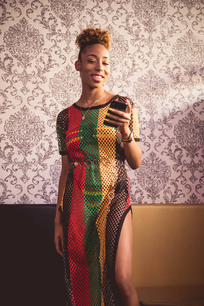 Vertical photo of the portrait of a black girl with her hair tied up, big earrings and a colorful net dress smiling while using her mobile in front of a vintage wall - Photo, Image