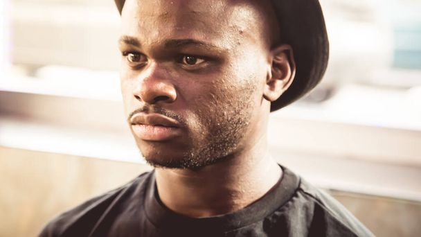 Πορτραίτο ενός μαύρου αγοριού σε ένα καπέλο που κοιτάζει ευθεία με μια σοβαρή έκφραση με ένα παράθυρο στο πίσω μέρος που τον φωτίζει - Φωτογραφία, εικόνα