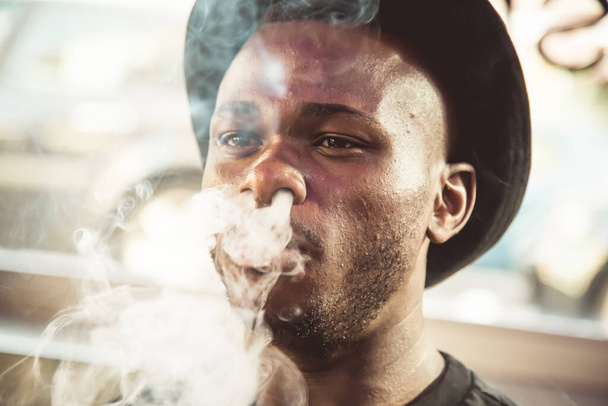 Portret van een zwarte jongeman met een hoed die rook uit zijn mond blaast in een kamer met een raam in zijn rug - Foto, afbeelding