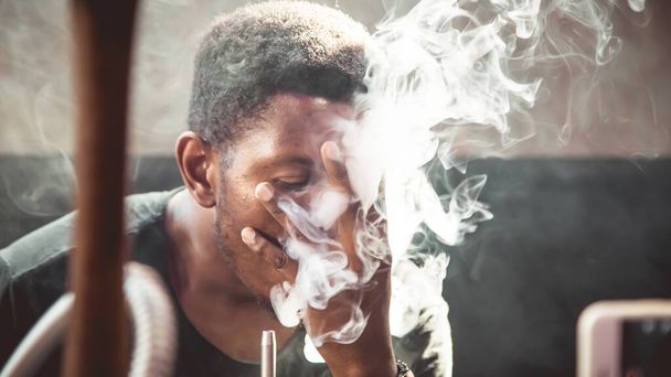 Πορτρέτο ενός μαύρου νεαρού άνδρα που καπνίζει ενώ αγγίζει το πρόσωπό του με το χέρι της και κάθεται σε καναπέ μέσα σε ένα δωμάτιο - Φωτογραφία, εικόνα