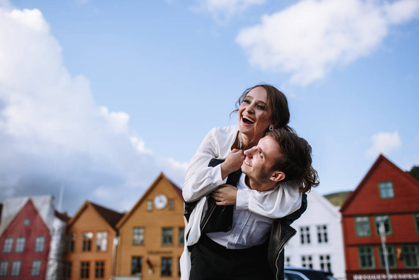 Two lovers against the background of the old quarter in the city of Bergen. The girl cheerfully climbed on the guy's back, people are smiling - Fotó, kép