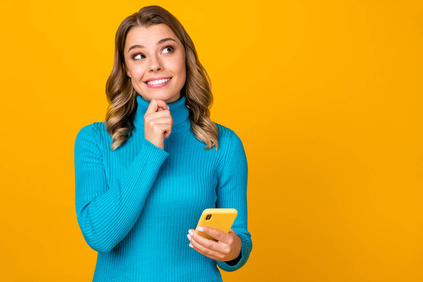 Photo of attractive curly lady blogger hold telephone writing new youth theme post look up empty space inspiration wear casual blue warm turtleneck isolated yellow color background - Foto, Imagen