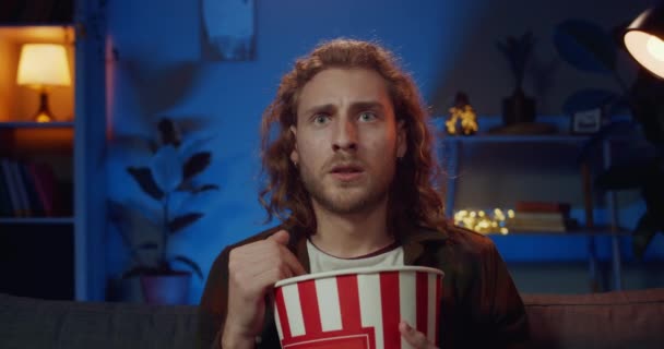 Young guy with long hair reacting on scary momment while watching thriller movie at home. Handsome man holding popcorn bowl while sitting on couch in front of tv. Concept of emotions. - Video