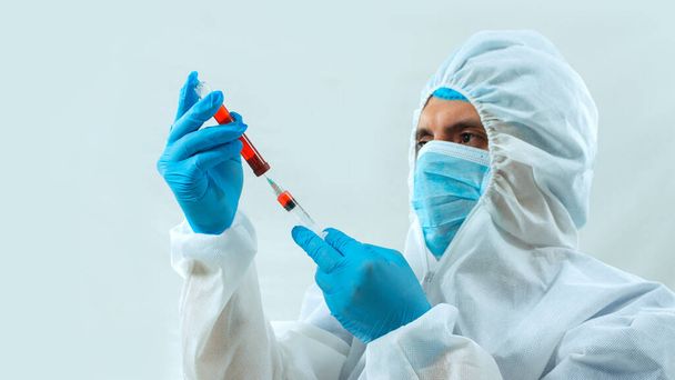 Latin doctor in bioprotective suit, face mask and blue gloves taking a blood sample in a test tube with a syringe on white background - Foto, immagini