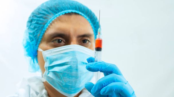Close-up zicht van de Latijnse arts in bioprotectief pak, gezichtsmasker en blauwe handschoenen starend met zijn ogen naar een spuit met een bloedmonster dat hij vasthoudt met zijn hand op witte achtergrond - Foto, afbeelding