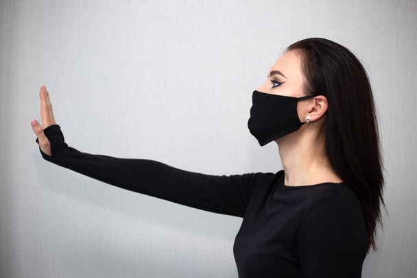 Studio portrait of young sexy woman wearing a surgical face mask and black clothes with outstretched arm on gray background. Flu and coronavirus epidemic, dust allergy, protection against virus. Beautiful social distance concept. Stop sign, - Photo, Image