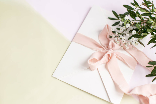 Φάκελος σε λευκό-ροζ φόντο με μεταξωτή κορδέλα ροδάκινο, πράσινο κλαδί και λουλούδια. Πρόσκληση γάμου. Κάρτα ημέρας της μητέρας. Θέση για κείμενο. Αντιγραφή χώρου. - Φωτογραφία, εικόνα