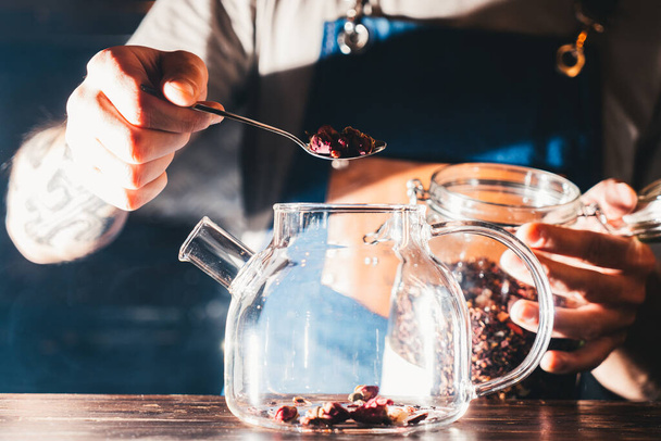 Barista is making rose herbal tea, filling glass teapot with dry rosebuds in a dramatic light of a bar, surrounded by light smoke; close up, hands visible - Photo, image