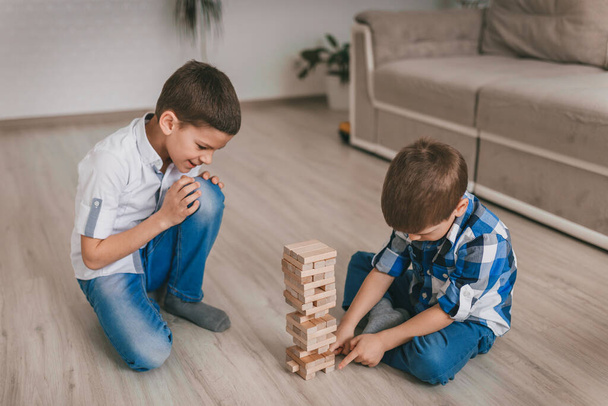 2人の兄弟の男の子が床に家の木製のブロックの塔を構築します。テーブルゲーム。独立した家族の隔離、感染症、コロナウイルス、自己分離の概念 - 写真・画像