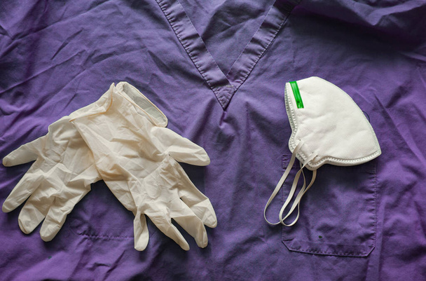 Gezichtsmasker en disposible handschoenen op paarse medische scrubs, Coronavirus Covid-19 concept achtergrond afbeelding - Foto, afbeelding