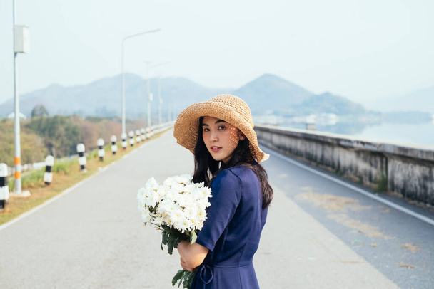 Una hermosa chica tailandesa parada en el camino de la presa sosteniendo las flores blancas en sus brazos volviendo a la cámara
. - Foto, Imagen