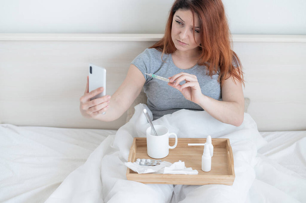 A cold blogger is recording a video for his vlog. A woman with a fever sits in bed and shows a thermometer to the smartphone camera. The girl is sick and takes a selfie. - Photo, image