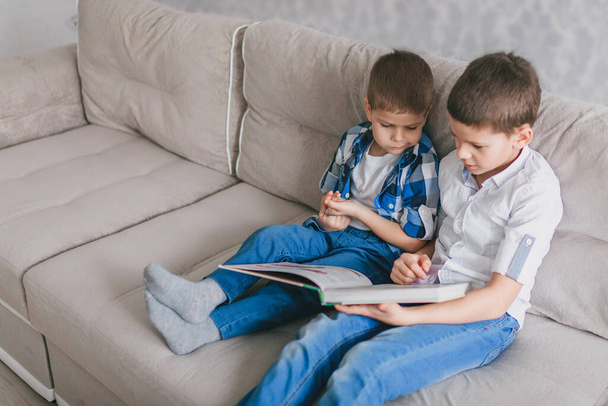 two brothers reading a book while sitting on a sofa in the living room during quarantine. The concept of social distance, self-isolation and home schooling - Zdjęcie, obraz