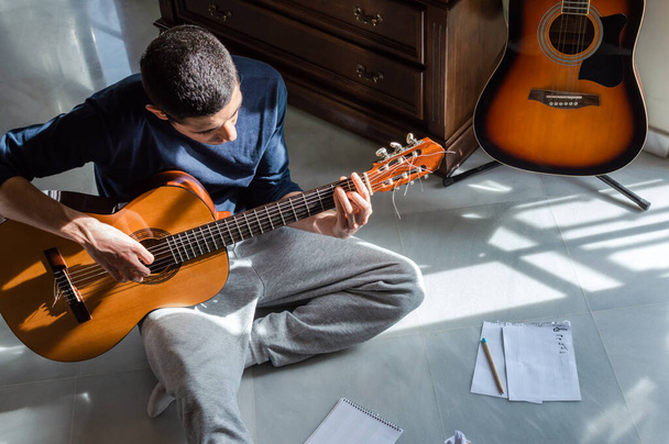 Man die thuis gitaar speelt en muziek componeert bij een helder raam op een zonnige dag. Casual muzikant die op de vloer zit en gitaar speelt. - Foto, afbeelding