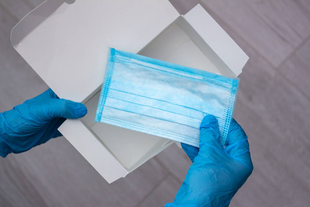 Hands wearing blue medical latex gloves hold an empty box and the last disposable protective medical mask, close-up, top view. Coronavirus pandemic concept - 写真・画像