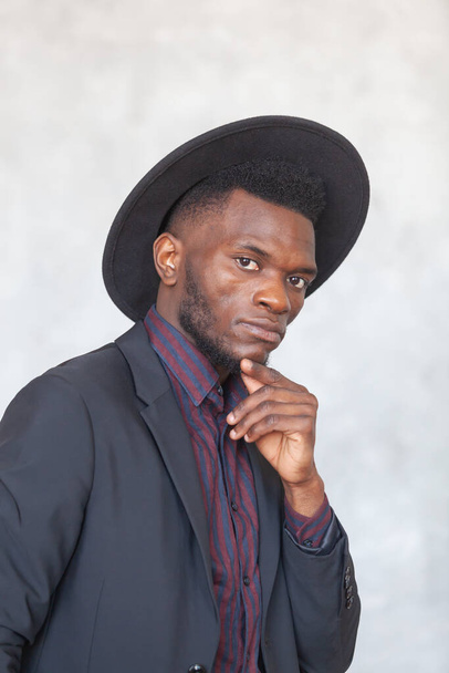 Young serious african man in a black suit and a hat looking at camera against a gray concrete wall - Fotoğraf, Görsel