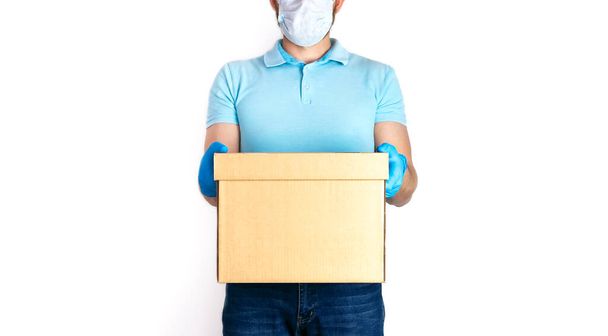 Concept of delivery of goods during quarantine 2019-ncov. Young man with beard in medical mask and blue gloves holds cardboard box in his hands on white background. Contactless delivery virus protect. - Photo, Image