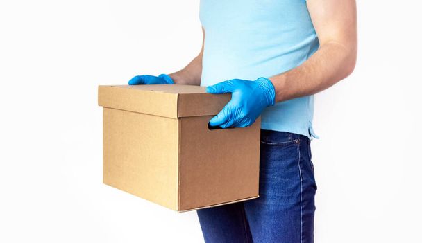 Concept van levering van goederen tijdens quarantaine. Jongeman in blauwe handschoenen houdt kartonnen doos in zijn handen op een witte achtergrond. Levering tegen Coronavirus 2019-nCov bij pandemie. Contactloze levering. - Foto, afbeelding