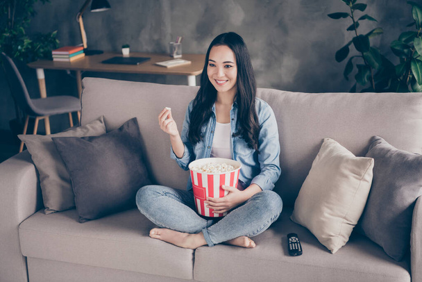 Portrait of candid dreamy cute girl sit divan legs crossed folded hold big pop corn box watch interesting movie in apartment indoors - Photo, Image