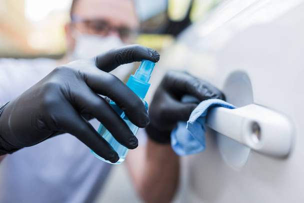 Driver with face mask wearing rubber protective gloves wiping and spraying car door handle. Virus and bacteria prevention concept. - Photo, image