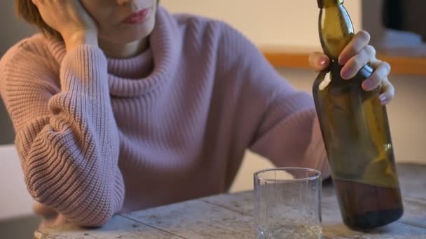 Sad woman in jeans and pink sweater is sitting at the table and drinking a whisky from the glass, dark green bottle is visible, female alcoholism - Footage, Video