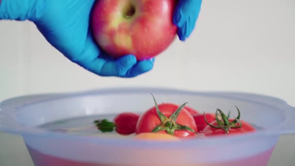 Mavi lateks eldivenli bir el mavi plastik bir kasede kırmızı bir elmayı suyla yıkıyor ve mutfakta çamaşır suyu var. Koronavirüsün yayılmasını önlemek için meyveyi dezenfekte ediyorum. - Video, Çekim
