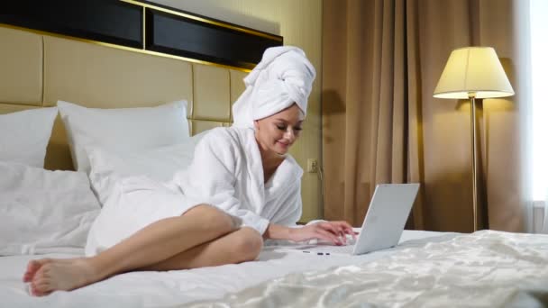 Młoda kobieta w białym szlafroku z ręcznikiem na głowie w domu z powodu kwarantanny pandemii koronowirusa COVID-2019. Praca zdalna lub odległa na laptopie w domu lub w apartamencie hotelowym. Materiał 4 k - Materiał filmowy, wideo