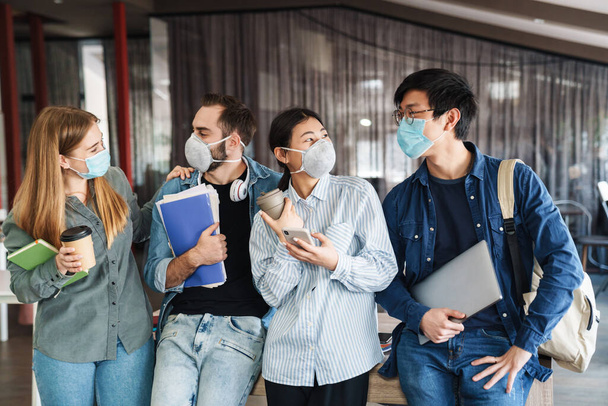 Photo of multinational joyful students in medical masks talking while standing at classroom - Foto, Imagen