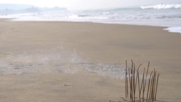 In de vroege ochtend lopen mannenvoeten langs het strand, aan de rand van het water, tegen de achtergrond van brandende wierookstokjes. Mooie rook. wazig achtergrond en ontwerp met kopieerruimte. - Video