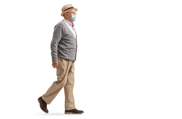 Full length profile shot of an eldelry man walking and wearing a protective medical face mask isolated on white background - Foto, imagen