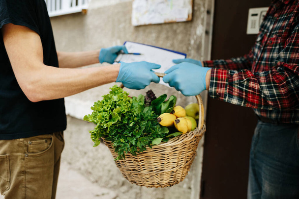 Delivery man wearing mask and gloves holding box basket full of different vegetables and greens. Customer receiving order from courier at home, organic fresh food online - Fotografie, Obrázek