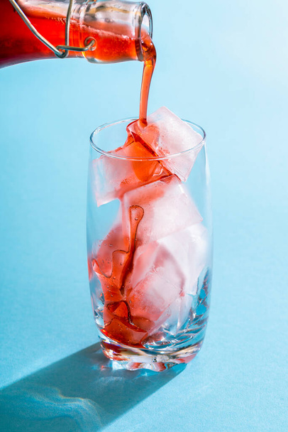Pouring strawberries syrup in a glass full of ice cubes. Homemade strawberry syrup made with organic fruits. Pouring red fruits syrup over ice cubes against a blue background. - Photo, Image