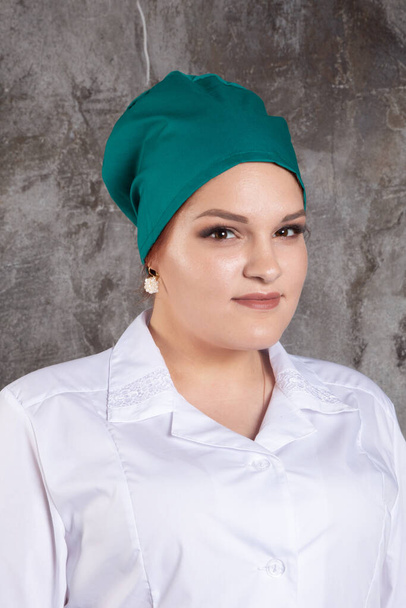 Portrait of attractive young female doctor in white medical jacket and  green medical cap on a grey background. - Foto, afbeelding