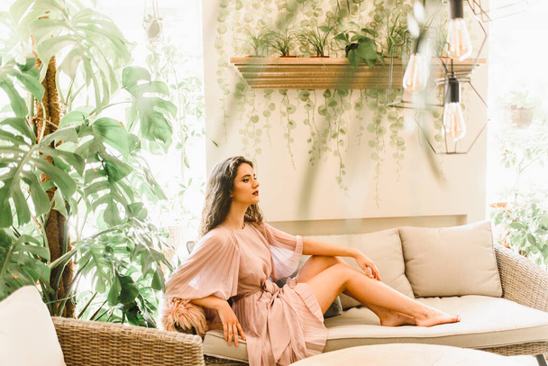 Joven mujer de negocios con el pelo ondulado en vestido rosa está sentado en el sofá suave en la cafetería. Mujer y plantas verdes - estilo ecologyl. Retrato de belleza
 - Foto, Imagen