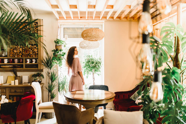 Professional model is standing opposite to window and looking to the right side. Natural beauty women with long wavy hair. Cafe with big green plants and palm tree - Fotoğraf, Görsel
