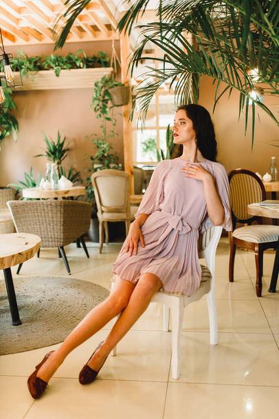 A fiatal nő puha rózsaszín ruhában ül a széken, és a bal oldalra néz. Szépség lány kávézó nagy zöld növények - Fotó, kép
