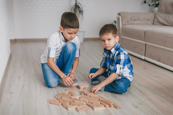 2人の兄弟の男の子が床に家の木製のブロックの塔を構築します。テーブルゲーム。独立した家族の隔離、感染症、コロナウイルス、自己分離の概念. - 写真・画像