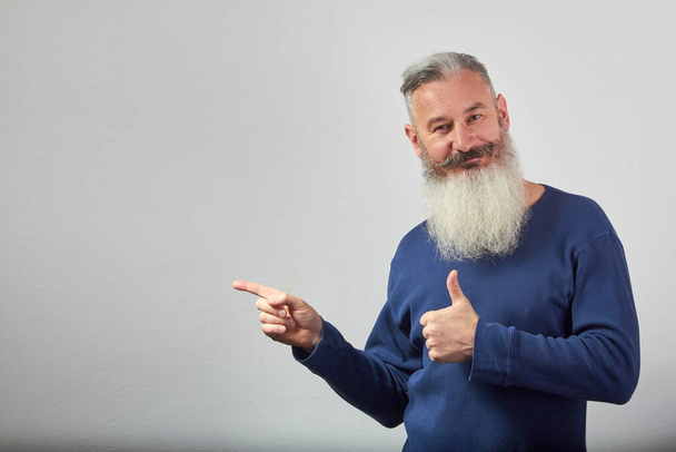 Portrait of mature gray-haired bearded man points forefinger to side and shows thumb up on grey background, selective focus - Фото, зображення