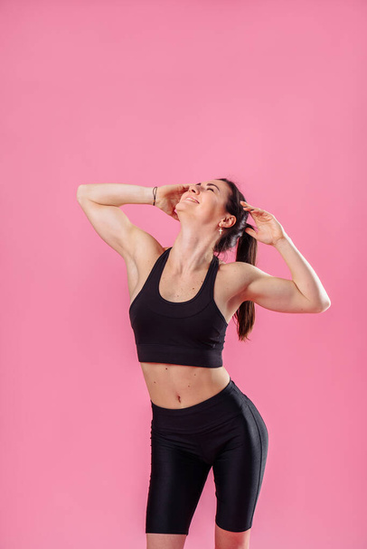 Attractive muscular girl does morning exercises, stretching sweetly, dreaming and smiling. Showing training movements in the studio, isolated on a pink background, wearing black top and pans. - Photo, Image