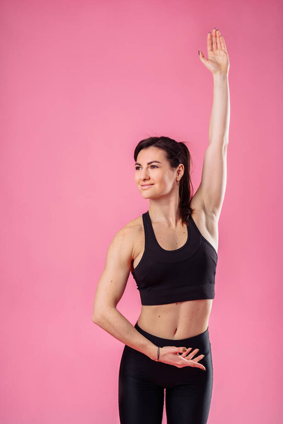 Attractive muscular girl does morning exercises, stretching sweetly, dreaming and smiling. Showing training movements in the studio, isolated on a pink background, wearing black top and pans. - Zdjęcie, obraz