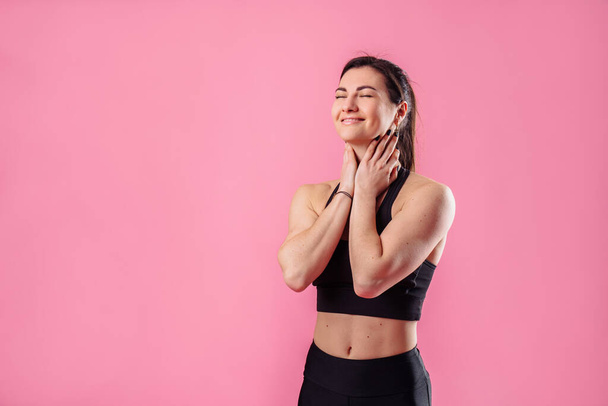 Тонкая накачанная девушка тренер фитнес стоит на розовом фоне в студии в черной спортивной одежде. Она обнимает себя руками, закрывает глаза, улыбается, мечтает и поднимает голову
 - Фото, изображение