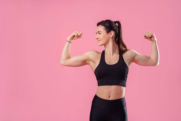 Half lenght portrait of beautiful, pumped up girl in a black tight-fitting top and leggings smiling and showing her big, pumped up muscles in her arms, isolated on pink background in photostudio - Photo, image