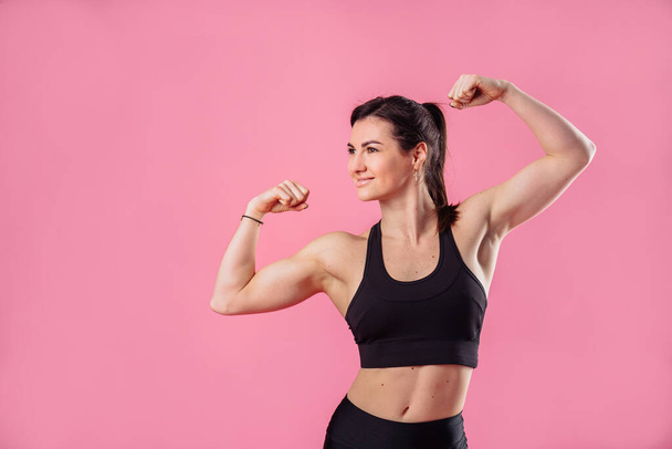 Half lenght portrait of beautiful, pumped up girl in a black tight-fitting top and leggings smiling and showing her big, pumped up muscles in her arms, isolated on pink background in photostudio - 写真・画像